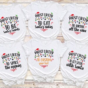 Op de afbeelding: Zes witte T-shirts met zwarte tekst en kleurrijke kerstverlichting. De tekst op de shirts luidt "Most Likely To" gevolgd door een andere kerstzin op elk shirt. De zinnen zijn: "Be Santa's Favorite", "Eat Santa's Cookies", "Drink All The Wine", "Spike The Eggnog", "Custom Your Text", en "Get Sassy With Santa".