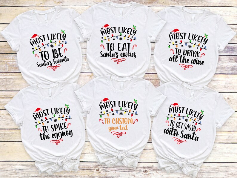 Op de afbeelding: Zes witte T-shirts met zwarte tekst en kleurrijke kerstverlichting. De tekst op de shirts luidt "Most Likely To" gevolgd door een andere kerstzin op elk shirt. De zinnen zijn: "Be Santa's Favorite", "Eat Santa's Cookies", "Drink All The Wine", "Spike The Eggnog", "Custom Your Text", en "Get Sassy With Santa".