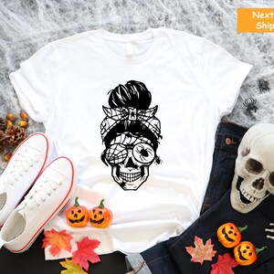 May include: White t-shirt featuring a black and white graphic of a skull with a spider web eye and a messy bun. The skull is wearing a bandana with a spider on it.