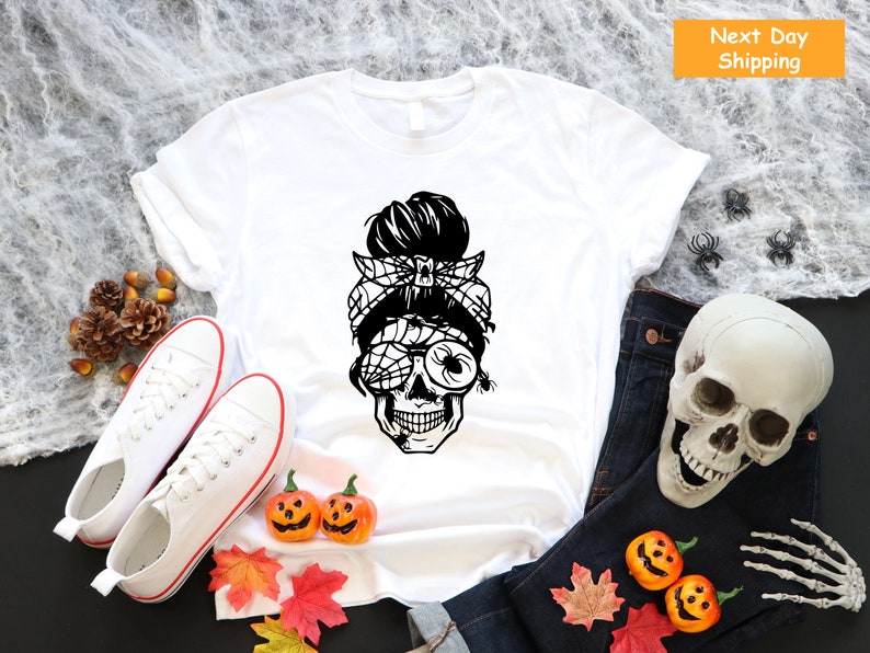 May include: White t-shirt featuring a black and white graphic of a skull with a spider web eye and a messy bun. The skull is wearing a bandana with a spider on it.