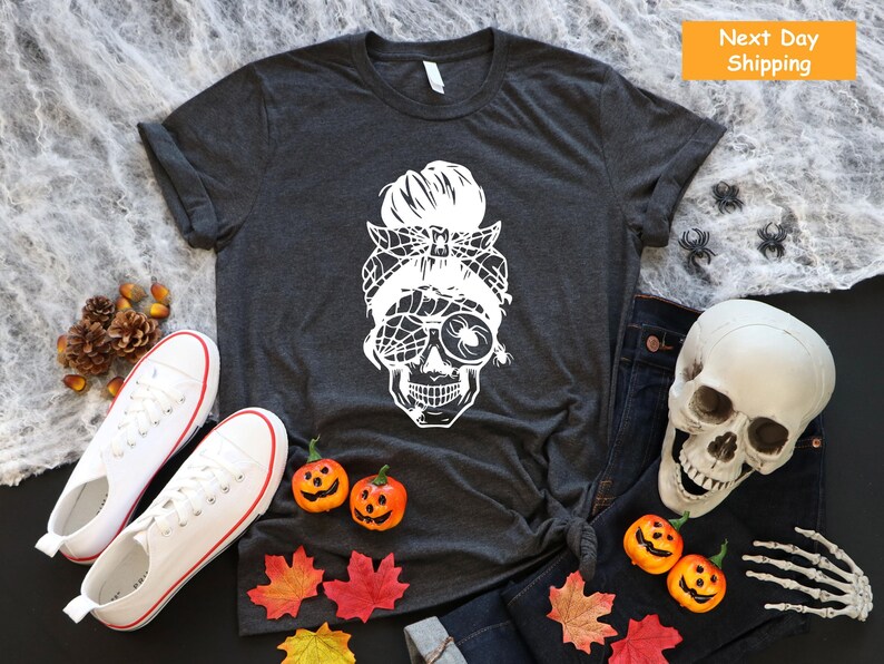 May include: A dark grey t-shirt with a white graphic of a skull with a messy bun and spider web accents. The graphic is centred on the shirt.