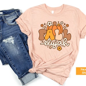 May include: A pale pink t-shirt featuring the word "FALL" in brown and orange, with a pumpkin graphic and the name "elyjah". The shirt is styled with blue denim jeans.