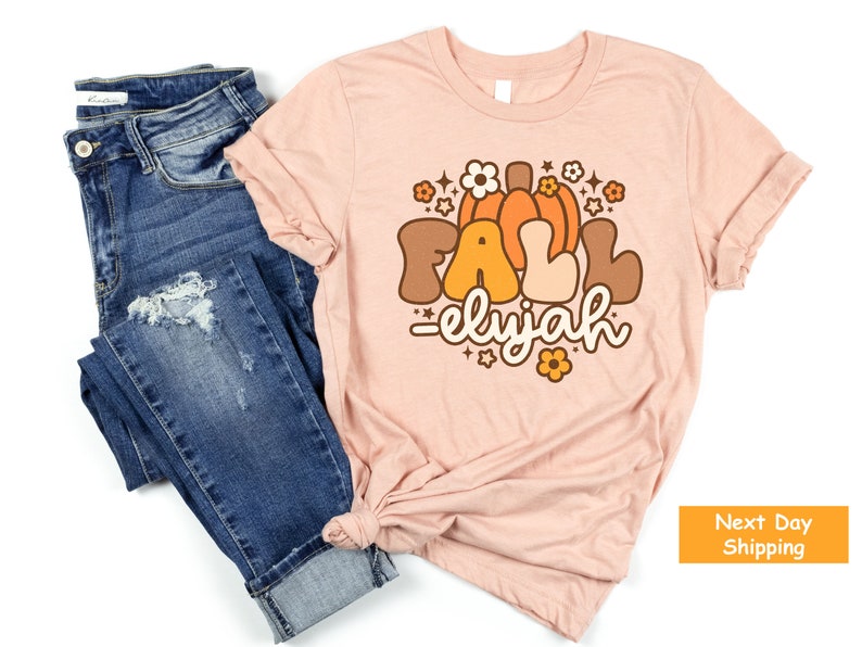 May include: A pale pink t-shirt featuring the word "FALL" in brown and orange, with a pumpkin graphic and the name "elyjah". The shirt is styled with blue denim jeans.