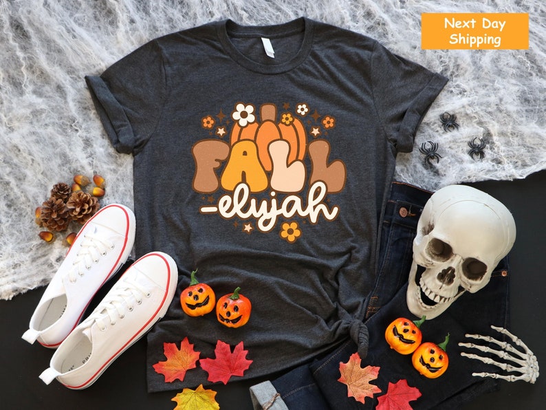 May include: A dark grey t-shirt featuring a retro "FALL - elijah" design in brown, orange, and white. The image includes white trainers, pumpkins, leaves, a skull, and spiders, suggesting a fall or Halloween theme.