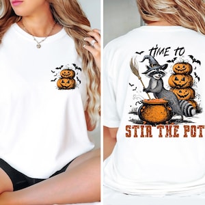May include: White t-shirt featuring a raccoon wearing a witch's hat and holding a broom, stirring a cauldron. The text "Time to Stir the Pot" is printed on the back of the shirt. The front of the shirt has a small image of three stacked pumpkins.