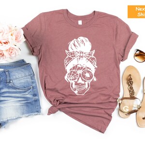 May include: A mauve t-shirt featuring a white graphic of a skull wearing sunglasses and a spider web headband. The skull has a messy bun hairstyle.