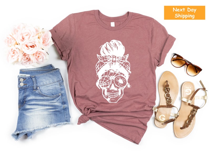 May include: A mauve t-shirt featuring a white graphic of a skull wearing sunglasses and a spider web headband. The skull has a messy bun hairstyle.
