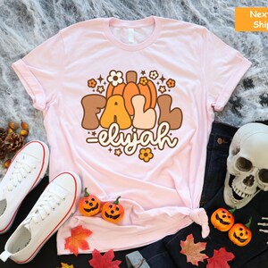 May include: A pale pink t-shirt featuring a retro graphic with the words "FALL elijah", pumpkins, and flowers. The shirt is styled with white trainers, a pair of jeans, and Halloween decorations.