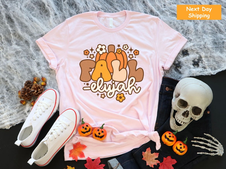 May include: A pale pink t-shirt featuring a retro graphic with the words "FALL elijah", pumpkins, and flowers. The shirt is styled with white trainers, a pair of jeans, and Halloween decorations.