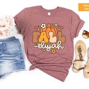May include: Mauve t-shirt featuring a retro graphic with the words "FALL -elyjah" and a pumpkin and floral design. The shirt is styled with denim shorts, sandals, and sunglasses, suggesting an autumn theme.