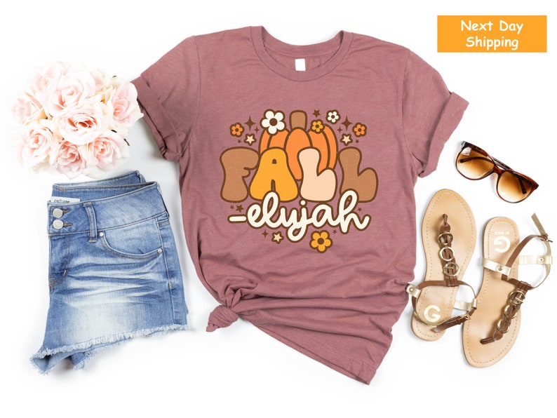 May include: Mauve t-shirt featuring a retro graphic with the words "FALL -elyjah" and a pumpkin and floral design. The shirt is styled with denim shorts, sandals, and sunglasses, suggesting an autumn theme.