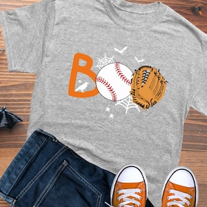May include: Gray t-shirt with an orange "BOO" graphic featuring a baseball, a baseball glove, and bats. The graphic also includes a spider web and a small bird.