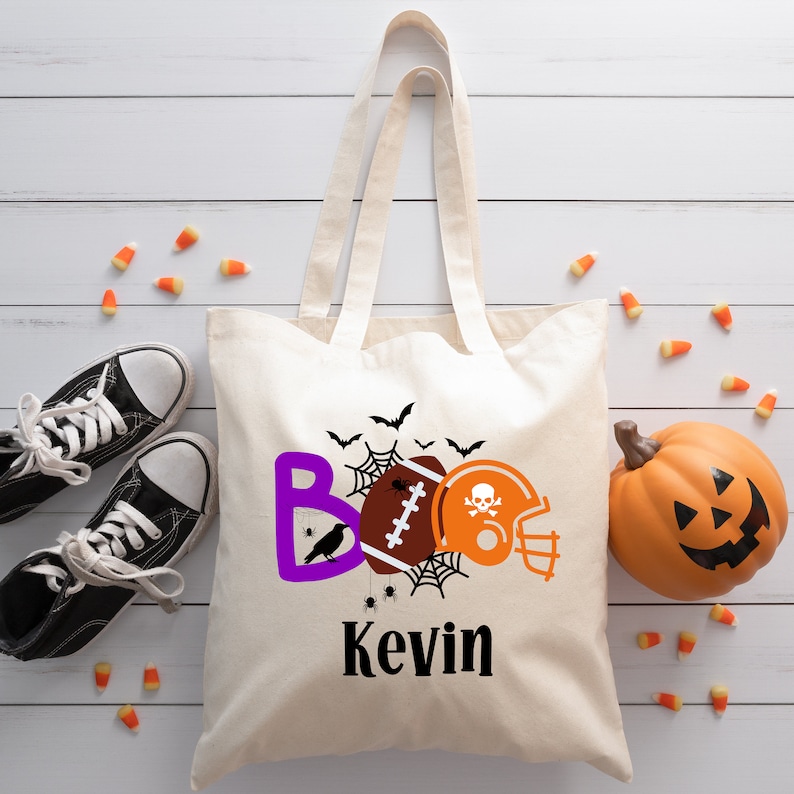 Op de afbeelding: Een witte canvas draagtas met een paars, oranje en zwart ontwerp met een voetbal, een helm, een spinnenweb en vleermuizen. De tekst "BOO" is paars en de naam "Kevin" is zwart.