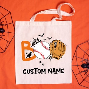 Op de afbeelding: Een witte canvas draagtas met een baseball, een honkbalhandschoen en een spinnenwebontwerp. De tekst "BOO" is oranje en de tekst "CUSTOM NAME" is zwart.