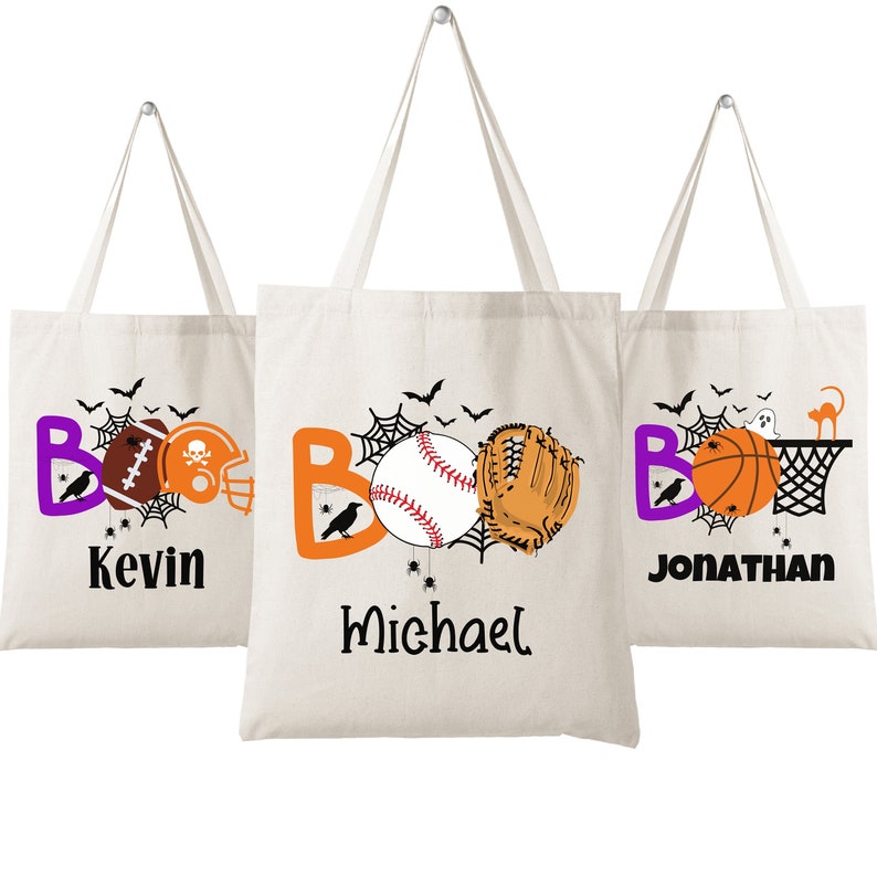 Op de afbeelding: Drie witte canvas tassen met het woord "BOO" in oranje en paarse letters. Elke tas heeft een ander sportthema: een voetbal en helm, een honkbal en handschoen, en een basketbal en ring. De tassen zijn gepersonaliseerd met de namen "Kevin", "Michael" en "Jonathan".