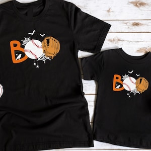 May include: Two black t-shirts with a baseball and glove design that says "BOO". The shirts are on a white wood background.