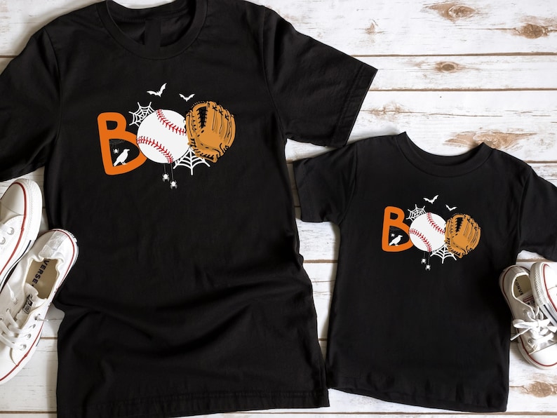 May include: Two black t-shirts with a baseball and glove design that says "BOO". The shirts are on a white wood background.