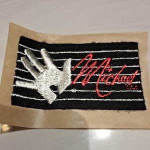 May include: Embroidered patch featuring a white hand gesture over a black and white striped background. The name "Michael" is written in red cursive script. The patch is on a brown paper backing.