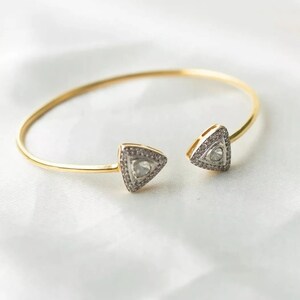 Puede incluir: Un brazalete de oro con dos dijes triangulares engastados con piedras transparentes. Los dijes están abiertos y el brazalete está abierto.