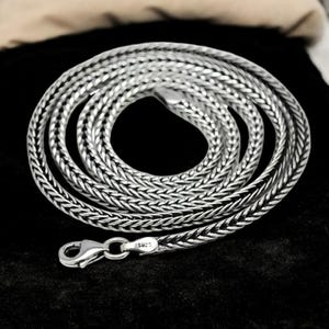 Op de afbeelding: Een zilverkleurige ketting met een gevlochten ontwerp. De ketting is gemaakt van een glanzend metaal en heeft een karabijnsluiting. De sluiting is gemarkeerd met "S925". De ketting wordt gepresenteerd op een zwarte ondergrond.