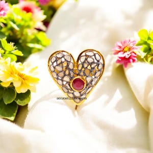 May include: A heart-shaped gold-toned ring with a central ruby and a border of small, irregularly shaped stones. The ring is set against a white background with colorful flowers in the background. The text "DEVJEWELLERSCRAFTS" is visible.