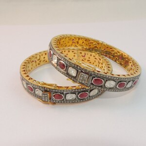Pode incluir: Dois braceletes de ouro com designs intrincados, adornados com pedras preciosas vermelhas e brancas. Os braceletes estão empilhados um em cima do outro.