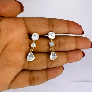 Pode incluir: Um par de brincos de ouro com três diamantes transparentes e facetados em cada brinco. O diamante superior é quadrado, o diamante do meio é redondo e o diamante inferior é em forma de pêra.