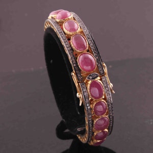 May include: A gold bangle bracelet featuring a row of oval-shaped pink gemstones set in a silver metal frame. The bracelet is adorned with small black diamonds.