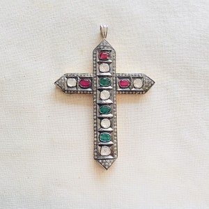May include: A silver cross pendant with a detailed design. The cross is adorned with green, red, and white gemstones.