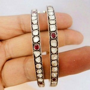 Pode incluir: Dois braceletes de prata com uma fila de pequenas pedras brancas e um acento de pedra vermelha em cada bracelete.