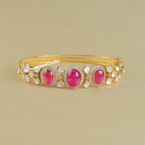 Puede incluir: Un brazalete de oro con tres piedras de rubí ovaladas engastadas en un lecho de diamantes blancos. El brazalete está hecho de oro y tiene un diseño delicado.