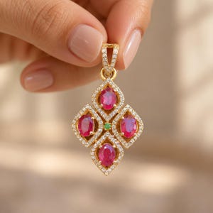 Puede incluir: Un colgante de oro con cuatro rubíes rojos de talla ovalada dispuestos en forma de diamante, rodeados de pequeños diamantes blancos en pavé. Una pequeña piedra preciosa verde está engastada en el centro y el asa está incrustada con diamantes.