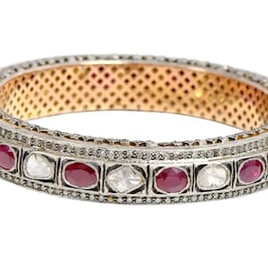 Pode incluir: Uma pulseira de prata e ouro com pedras preciosas vermelhas e transparentes alternadas. A pulseira apresenta uma fileira de pequenos diamantes e um interior dourado. As pedras preciosas são engastadas numa moldura prateada.