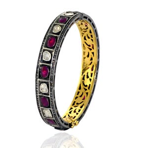 May include: A silver bangle bracelet with a gold interior. The bracelet is decorated with a row of red gemstones and white diamonds.