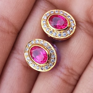 May include: A pair of gold earrings with oval-shaped red gemstones surrounded by a halo of smaller white gemstones.