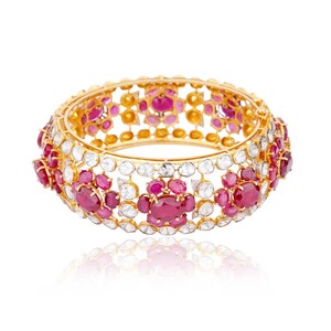 May include: A gold bangle bracelet with a intricate design featuring red and white gemstones. The bracelet is set against a white background.