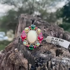 May include: A silver ring with a large white opal surrounded by smaller red, green, and yellow gemstones. The ring is on a wooden surface. The tag reads "SG 5D Silver P: 2700".