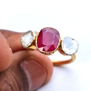 Può includere: Un anello d'oro con tre pietre. La pietra centrale è un grande rubino rosso. Le due pietre ai lati sono diamanti bianchi più piccoli.