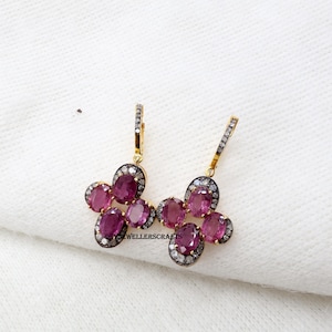 May include: Gold-coloured drop earrings with clusters of oval, deep pink gemstones. Each cluster is encircled by a halo of small, sparkling stones. The earrings are set against a white background.