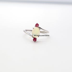 May include: A silver ring featuring an oval, off-white opal gemstone, flanked by two small, round, red gemstones. The ring has a delicate, minimalist design. The text "DEVJEWELLERSCRAFTS" is visible.