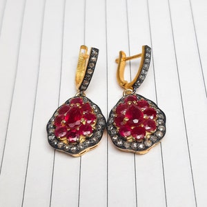 Puede incluir: Un par de pendientes de oro con un racimo de piedras de rubí rojo rodeadas de piedras transparentes más pequeñas. Los pendientes tienen un diseño colgante.