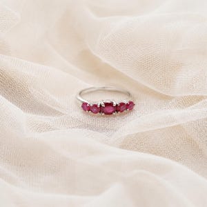May include: A silver ring featuring five prong-set, round-cut red rubies arranged in a horizontal row. The center ruby is slightly larger than the four flanking stones, creating a graduated design on a polished, thin, circular metal band.