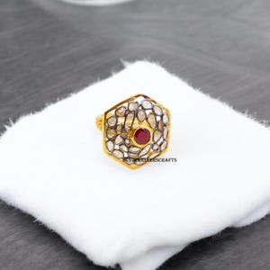 Op de afbeelding: Een gouden ring met een zeshoekig ontwerp met een rode edelsteen omringd door kleinere witte edelstenen. De ring ligt op een witte achtergrond. DEVJI JEWELLERSCRAFTS is gedrukt op de achtergrond.