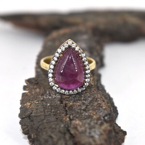 May include: A gold ring featuring a teardrop-shaped purple gemstone encircled by a halo of small white gemstones.