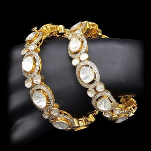 Op de afbeelding: Twee gouden armbanden met witte edelstenen. De armbanden zijn versierd met ingewikkelde ontwerpen en bezet met ronde en ovale stenen.