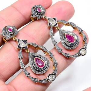 May include: A pair of dangle earrings with a teardrop-shaped ruby gemstone surrounded by smaller diamonds. The earrings are made of silver and gold and have a delicate, intricate design.