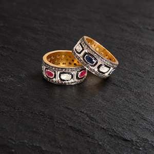 May include: Two gold bands featuring rectangular settings with embedded red rubies, blue sapphires, and clear stones. The exterior surfaces are encrusted with small pavé diamonds set in dark metal, contrasting with the polished gold interior finish.