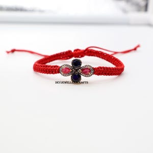 May include: Red macrame bracelet featuring a four-petal floral design. The flower has ruby and sapphire gemstones, with a silver-colored metal setting. The bracelet is adjustable with a sliding knot closure. The text "DEVJEWELLERCRAFTS" is visible.