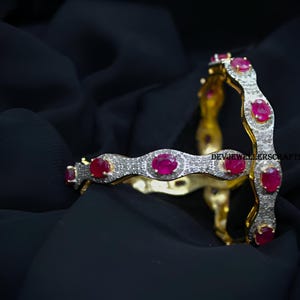 Könnte beinhalten: Zwei goldene Armreifen mit einem wellenförmigen Design, besetzt mit kleinen klaren Steinen und größeren roten ovalen Steinen. Die Armreifen sind übereinander gestapelt.