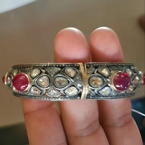 以下が含まれることがあります： 黒のエナメルデザインと赤い宝石が施された銀と金のバングルブレスレット。ブレスレットは小さなダイヤモンドで飾られています。